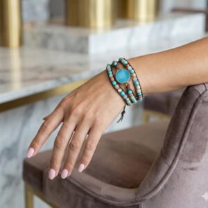 Woman's hand resting on a marble table wearing a blue druzy and turquoise wrap bracelet. The setting features chic gold accents and a velvet chair, showcasing a luxury boho look.