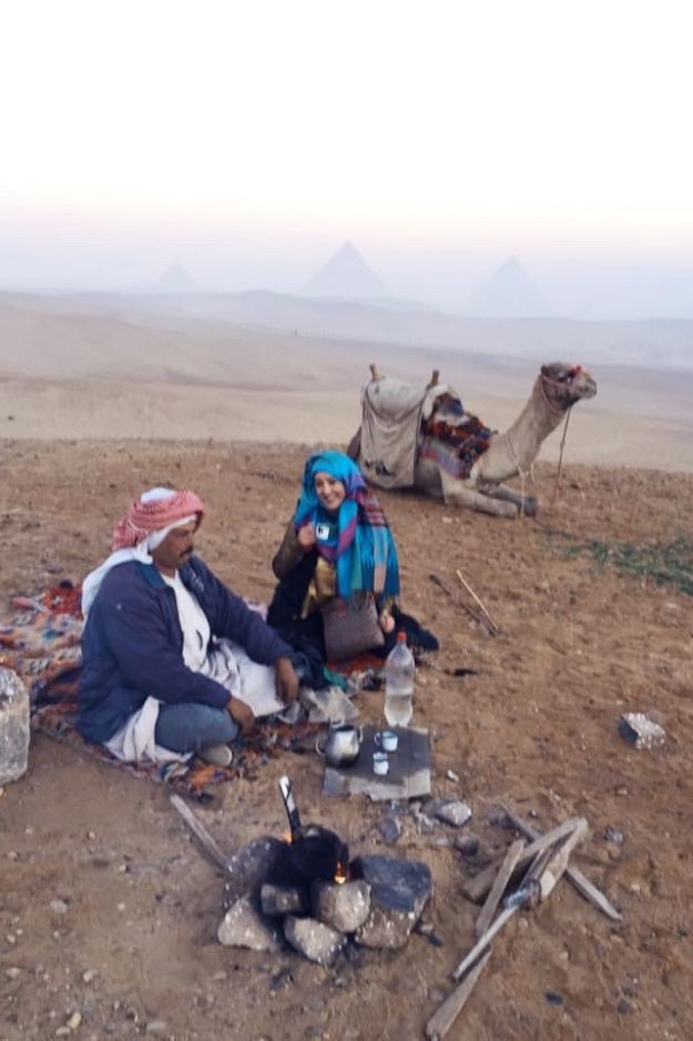 A woman and a local sitting in the Egyptian desert near the Pyramids of Giza, sharing a traditional experience with a camel resting nearby.