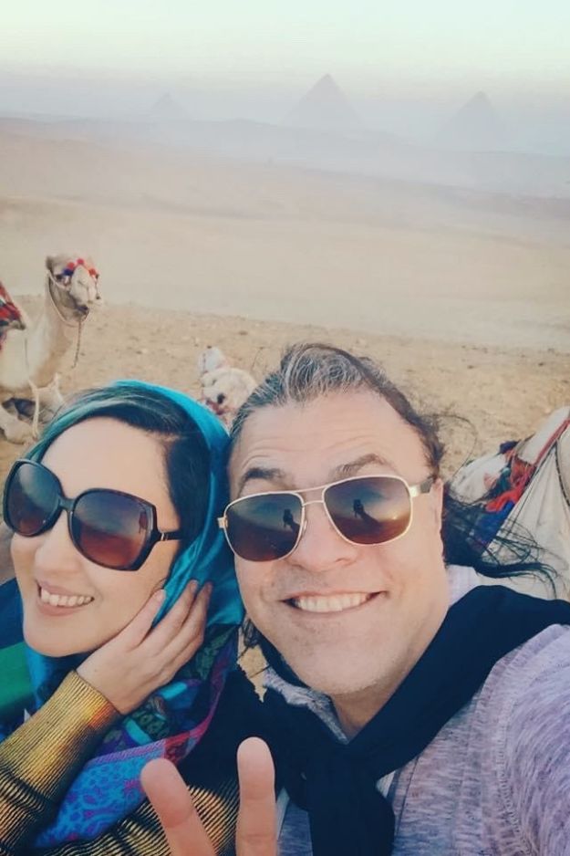 A couple taking a selfie in the Egyptian desert with the Pyramids of Giza in the background and camels nearby, enjoying a memorable adventure.