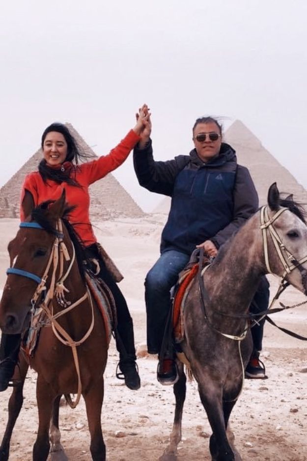 A couple on horseback raising their hands together in front of the Pyramids of Giza, Egypt, enjoying a memorable adventure.