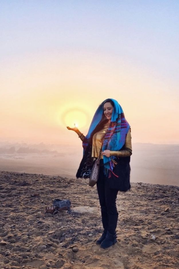 A woman standing in the desert near the Pyramids of Giza, appearing to hold the setting sun in her hand, capturing a magical moment in Egypt.