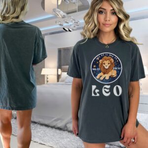 Woman wearing a Pepper Comfort Colors 1717 t-shirt with a Leo Zodiac design featuring a cute lion illustration, stars, and the dates "July 23 to Aug 22."